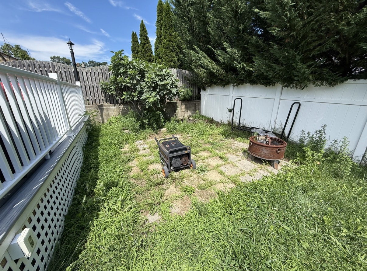 Weedy backyard beside a deck and white fence before sod installation